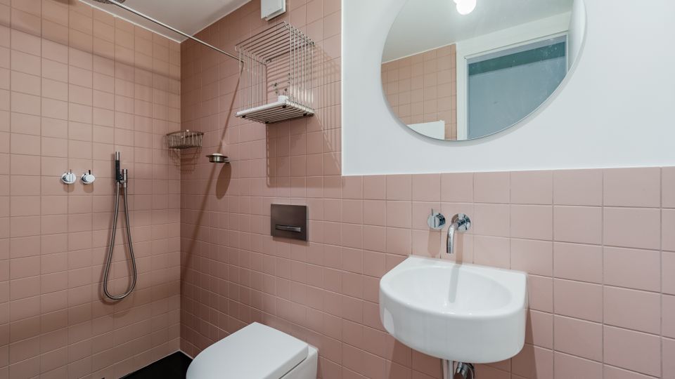 Kleines Badezimmer mit rosa Wandfliesen, bodengleicher Dusche mit Regenduschkopf und Handbrause sowie wandhängendem WC. Rechts sind ein kleines Waschbecken mit Wandarmatur und ein runder Spiegel über der halbhoch gefliesten Wand zu sehen.