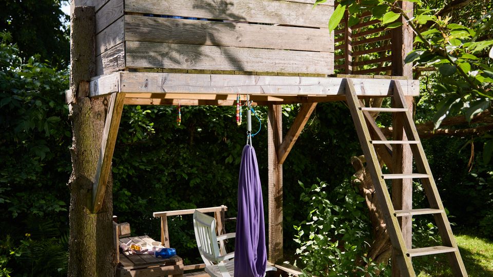Holz-Baumhaus auf Stelzen in einem dicht bewachsenen Garten, mit kleiner Veranda, seitlicher Leiter und blauem Vorhang am Fenster. Darunter befindet sich eine Holzplattform mit Sitzmöbeln und Sonnenschirm, umgeben von Bäumen und Sträuchern.