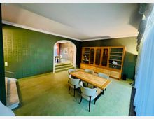 Wohnzimmer im klassischen Stil mit grüner Tapete und grünem Teppichboden. Im Vordergrund steht ein rechteckiger Esstisch mit sechs hell gepolsterten Stühlen, im Hintergrund ein großer Holzbücherschrank und ein Durchgang mit Rundbogen, der zu einem angrenzenden Raum führt.