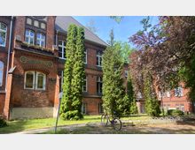 Backsteingebäude mit der Aufschrift "Kreis-Krankenhaus", umgeben von hohen Bäumen. Vor dem Gebäude steht ein Fahrrad an einem Fahrradständer, und der Rasenbereich ist von einem gepflasterten Weg umgeben.