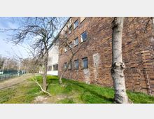 Backsteinfassade eines mehrstöckigen Gebäudes mit vergitterten Fenstern, dahinter eine Grünfläche mit kahlen Bäumen. Rechts neben dem Gebäude verläuft ein schmaler Wiesenstreifen, während links ein Zaun und ein Weg sichtbar sind.