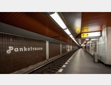 U-Bahn-Station mit dem Namen „Pankstraße“, dargestellt durch weiße Schrift auf einer braunen gefliesten Wand. Der Bahnsteig ist sauber und mit einem orangefarbenen Deckenpaneel sowie Beleuchtung ausgestattet; sichtbare Gleise verlaufen parallel zur Wand.