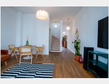 Wohnraum mit hellem Holzfußboden, einem runden Esstisch aus Holz und vier passenden Stühlen. Im Hintergrund führt eine Holztreppe in das obere Stockwerk, Wände sind weiß, Pflanzen in Töpfen und moderne Lampen betonen das minimalistische Design.