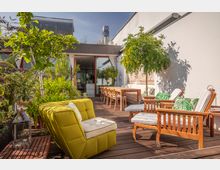 Stilvoll gestaltete Dachterrasse mit Holzboden, grünen Pflanzen und moderner Einrichtung, darunter ein gelber Sessel und Holzstühle mit Polstern. Im Hintergrund ein Esstisch mit Stühlen und eine Glasfassade, die zu einem Innenraum führt.