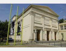Das Bild zeigt die Fassade des Maxim Gorki Theaters in Berlin, ein klassizistisches Gebäude mit Säulen und Reliefs. Vor dem Eingang stehen gelbe Fahnenmasten mit Plakaten, auf denen schwarz-weiße Theaterdarstellungen zu sehen sind.