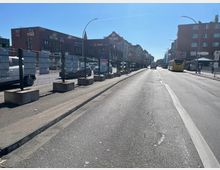 Eine Straße in einer städtischen Umgebung mit mehrspurigem Verkehr, flankiert von Gebäuden aus rotem Backstein und Geschäften wie "dm" und "Rewe". Auf der rechten Straßenseite befindet sich eine Bushaltestelle mit einem gelben Bus, während links ein Parkplatz hinter einem Metallzaun erkennbar ist.