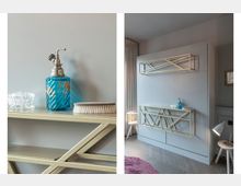 Modernes Innenraumdesign mit einer hellgrauen Wand, an der zwei geometrisch gestaltete, goldfarbene Regale montiert sind. Auf dem unteren Regal stehen eine blaue Glasflasche, eine Bürste und ein kleines Kristallgefäß; die Umgebung ist mit einem hellen Holzfußboden, einem weißen Beistelltisch mit Lampe und einem lilafarbenen Teppich gestaltet.