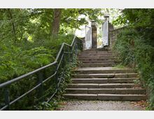 Steintreppe in einem von Bäumen und Sträuchern gesäumten Park, die zu einem offenen weißen Tor mit dekorativen Säulen führt. Die Umgebung ist grün und dicht bewachsen, mit einem Geländer an der Seite der Treppe.