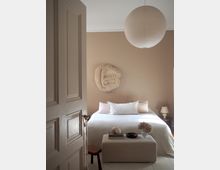 Minimalist bedroom with beige walls and a white bed, featuring a textured wall sculpture above the headboard and a large round paper pendant light on the ceiling. A low upholstered bench at the foot of the bed holds books and decor, with a small bedside table and lamp beside an open paneled door.