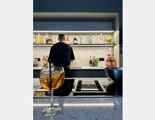 Ein modernes Kücheninterieur mit einer marmoranen Arbeitsfläche und Induktionskochfeld im Vordergrund. Im Hintergrund stehen ein Mann in schwarzer Kleidung vor einem beleuchteten Regal mit zahlreichen Spirituosenflaschen und Cocktailzutaten.