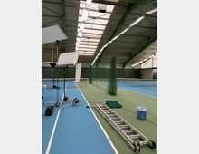 Eine Tennishalle mit grünen und blauen Tennisplätzen, hoher Decke mit Metallträgern und Beleuchtung. Im Vordergrund befinden sich eine Leiter, Scheinwerfer und technische Ausrüstung.