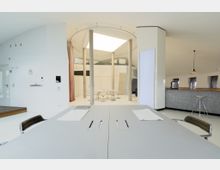 Moderner Innenraum mit minimalistischer Einrichtung, dominiert von weißen Wänden und klaren Strukturen. In der Mitte befindet sich ein runder Bereich mit Betonpfeilern und einem Glasdach, daneben eine Theke mit Barhockern sowie ein schlichter Konferenztisch im Vordergrund.