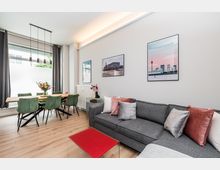 Heller, moderner Wohn- und Essbereich mit grauem Ecksofa und farbigen Kissen, rotem Couchtisch sowie Holzboden. Im Hintergrund steht ein Esstisch mit grünen Stühlen neben großen Fenstern mit Vorhängen; an der Wand hängen gerahmte Bilder und eine schlanke Hängelampe beleuchtet den Tisch.