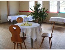 Ein schlichter Raum im Landhausstil mit Holzmöbeln. Runde und rechteckige Tische sind mit weißen Tischdecken und einfachen Blumengestecken dekoriert, die Stühle haben herzförmige Ausschnitte in der Rückenlehne. Große Fenster lassen Tageslicht herein, und eine Zimmerpflanze steht in der Ecke des Raumes.