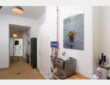 Ein heller Flur mit beigem Fliesenboden, einer Wand mit einem Bild von Sonnenblumen und einem Holztisch mit Kaffeemaschine, Tassen sowie Küchenutensilien. Im Hintergrund ein Raum mit Fotostudio-Ausrüstung wie Stativen und Beleuchtung.