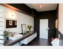 Modernes Badezimmer mit dunklen Wänden und weißen Akzenten, einschließlich eines Waschtischs mit rechteckigem Waschbecken, Spiegel und Pflanzen. Im Hintergrund befinden sich eine weiße Tür, eine Toilette und Wanddekoration mit Pflanzenmotiven.