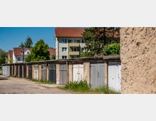Reihe älterer Garagen mit unterschiedlich gestrichenen Holztoren entlang einer asphaltierten Zufahrt, teils mit Bewuchs am Boden. Dahinter stehen mehrgeschossige Wohnhäuser mit roten Ziegeldächern und Bäumen unter blauem Himmel.