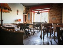 Gemütlicher Innenraum eines Cafés mit einer Backsteinwand, Holzboden und einer Einrichtung aus dunklen Ledersofas, Holzstühlen und Tischen. Große Fenster lassen Tageslicht herein, und eine Retro-Stehlampe sowie Wanddekorationen ergänzen das Ambiente.