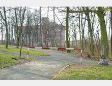 Ein mit Bäumen gesäumter Weg im Freien mit einer rot-weiß gestrichenen Barriere, die den Weg versperrt, im Hintergrund ein mehrstöckiges Backsteingebäude.