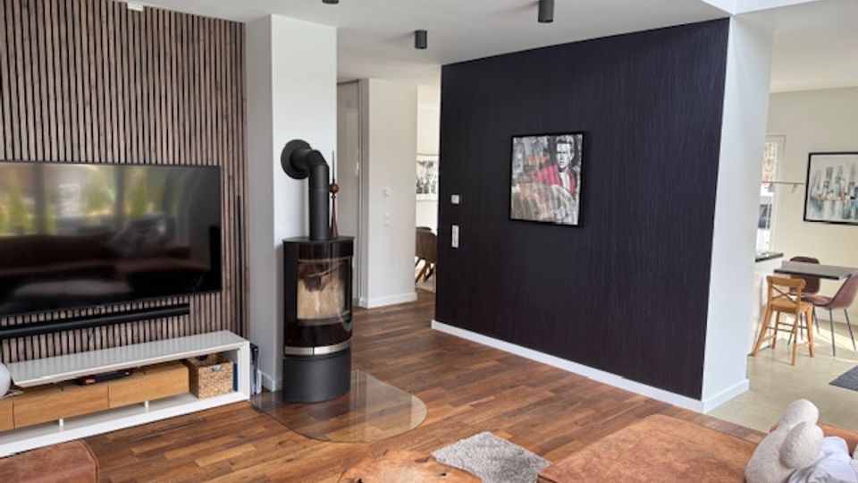 Alt text: Modern open-plan living room with wood floors, a wall-mounted TV on a slatted wood accent wall, and a black freestanding wood stove near a dark feature wall with framed art. The space opens into a bright dining area with light carpet and simple furniture.