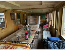 Alter Innenraum eines gelben, umgebauten Waggons mit langen Holztischen und Bänken. Der Raum ist mit Fenstern und gelben Wänden ausgestattet, die vereinzelt mit Bildern und Dekorationen versehen sind; Gegenstände wie Decken, Stühle und Haushaltsartikel sind verteilt.