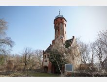 Historischer Backstein-Wasserturm mit achteckigem Schaft und Metallkuppel, an den ein kleines Gebäude mit Satteldach und Rundbogenportal anschließt. Der Turm steht in einer parkähnlichen Umgebung mit kahlen Bäumen und Sträuchern unter klarem Himmel.