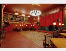 Theaterraum im Vintage-Stil mit rotem Teppichboden und einer Bühne, die durch einen Vorhang in kräftigem Rot verdeckt wird. Es gibt eine Galerie mit geschwungenem Geländer, eine Wendeltreppe, Kronleuchter und dekorative Wandverzierungen mit goldenen Details.