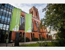 Backsteinbau mit hohen gotischen Fenstern und einem markanten Turm, genutzt als "Umweltforum", erkennbar durch grüne Banner mit der Aufschrift. Im Vordergrund ein grüner Gehweg mit Bäumen und Sträuchern.