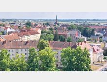 Stadtansicht mit Wohnhäusern in Fassadenreihe, roten Ziegeldächern und grünen Bäumen im Vordergrund, im Hintergrund eine Kirche mit spitzem Turm sowie Felder und Wiesen in der Ferne.