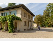Historisches Gebäude mit heller Fassade und Vorbau, bewachsen mit grünem Efeu sowie dekoriert mit Skulpturen über dem Eingang. Umgeben von gepflastertem Platz und Bäumen in einer parkähnlichen Umgebung.