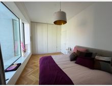 Schlafzimmer mit weiß gestrichenen Wänden, großem Fenster mit Fensterbank, Holzboden in Fischgrätmuster, großem Bett mit lilafarbener Decke und Kissen in verschiedenen Farben. Im Hintergrund ein weißer Einbauschrank und eine Tür.