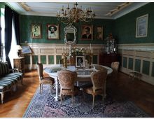 Ein elegantes, historisch eingerichtetes Zimmer mit grünen Tapeten, einem ovalen Tisch in der Mitte, umgeben von gepolsterten Stühlen mit Verzierungen. An den Wänden hängen gerahmte Porträts, ein Kronleuchter hängt von der Decke, und der Holzboden ist mit einem gemusterten Teppich bedeckt.