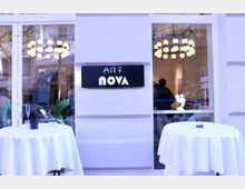 Fassade eines modernen Gebäudes mit großen Fenstern, hinter denen ein Ausstellungsraum sichtbar ist. Auf einem weißen Schild steht "Art Nova", davor stehen kleine Stehtische mit weißen Tischdecken.