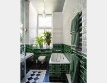 Kleines Badezimmer mit grünen glänzenden Fliesen an der unteren Wandhälfte und weißen Metrofliesen oben. Es gibt eine Badewanne, ein wandmontiertes WC, ein Waschbecken und ein Fenster, vor dem mehrere grüne Pflanzen stehen. Der Boden hat ein schwarz-weißes Fliesenmuster.