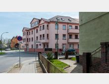 Eine Straßenecke in einer Wohngegend mit einem mehrstöckigen, hellrosa Wohnhaus mit roten Fensterrahmen und Schrägdach im Mittelpunkt. Im Vordergrund befinden sich ein Bürgersteig, ein Zaun, sowie mehrere Straßenschilder, darunter ein Halteverbotsschild und ein Wegweiser mit der Aufschrift „Am Ritterfeld“.