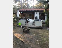 Kleines Gartenhaus mit Terracotta-Dach, davor eine Holzterrasse mit Korbmöbeln und einem Sonnenschirm. Im Garten steht eine Feuerstelle, und der Bereich ist von Bäumen umgeben.