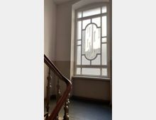 Treppenhaus mit einem großen, verzierten Fenster mit mattem Glas und Blick auf die Fassade eines gegenüberliegenden Gebäudes; im Vordergrund ein hölzernes Treppengeländer und ein dunkler Boden.