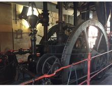 Innenansicht einer historischen Dampfmaschine in einer Fabrikhalle, mit sichtbarem Schwungrad, Kolben und Zahnrädern. Die Umgebung zeigt einen gealterten Industriebereich mit rohrartigen Strukturen und gedämpfter Beleuchtung.