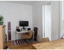 Ein einfach eingerichtetes Zimmer mit einem Schreibtisch, auf dem ein Monitor, Lautsprecher und ein Verstärker stehen. Im Vordergrund ein Holztisch mit einem Stuhl, ein gemusterter Teppich auf Holzboden, und eine offene Tür führt zu einem angrenzenden Raum.