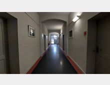 Langer, schlichter Flur in einem Gebäude mit hellen Wänden, schwarzen Böden und roten Randstreifen; an den Wänden hängen gerahmte Bilder, und am Ende des Flurs ist eine doppeltürige Glasfront mit Tageslicht sichtbar.