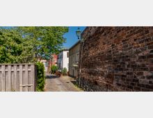 Schmale, gepflasterte Gasse mit kleinen Wohnhäusern und einer hohen Ziegelmauer auf der rechten Seite; links ein Holzzaun und dahinter grüne Bäume, oben eine Laterne im klassischen Stil.