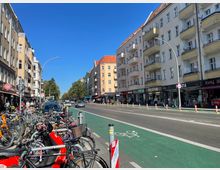 Straßenszene in einer Stadt mit mehrstöckigen Wohnhäusern auf beiden Seiten, Erdgeschosse mit Geschäften und Restaurants. Im Vordergrund ein Fahrradabstellbereich, daneben ein grüner Radweg, sowie eine Fahrbahn mit Autos und sichtbaren Fußgängern bei sonnigem Wetter.