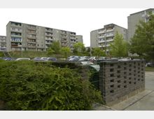 Mehrstöckige Wohnblöcke aus Beton mit Balkonen, umgeben von einem Parkplatz voller geparkter Autos. Im Vordergrund sind Büsche und eine Steinmauer mit einem kleinen, durchbrochenen Gittertor zu sehen, daneben einige Bäume.