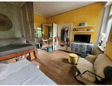 Gemütliches Zimmer mit Holzboden und gelben Wänden. Zu den Möbeln gehören ein Hochbett mit Vorhang, ein Sofa mit Fellüberwurf, ein Fernseher auf einer Kommode mit Stoffschubladen, sowie ein großer Wandspiegel, der den Raum optisch erweitert.