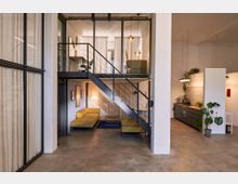 Offener Loft-Innenraum mit Betonboden, großer Glas-Trennwand und schwarzer Metalltreppe zu einer Galerieebene mit Vorhängen und Hängeleuchten. Unten befindet sich ein Sitzbereich mit Sofa, daneben eine moderne Küchenzeile mit Arbeitsplatte und Zimmerpflanzen.