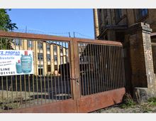 Geschlossenes rostiges Metalltor mit Stacheldraht oben, dahinter ein verlassenes Fabrikgebäude aus gelbem Backstein mit großen Fenstern. Im Vordergrund ein Schild, das Flüssiggaspreise und Kontaktdaten bewirbt.