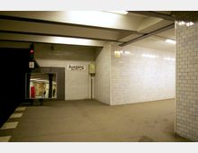 Unterirdischer U-Bahnhof mit weißen, gefliesten Wänden und einer Decke mit sichtbaren Trägern. Ein Schild mit der Aufschrift „Ausgang“ weist auf den Ausgang hin, und ein Spiegel reflektiert den Blick auf den gegenüberliegenden Bahnsteig mit wenigen Personen.