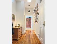 Langer, schmaler Flur mit Holzdielenboden in einer Wohnung. Links steht eine Holzkommode mit Dekoration, rechts hängen zahlreiche Bilderrahmen an der Wand, und am Ende des Flurs ist ein rustikaler Holzschrank zu sehen.