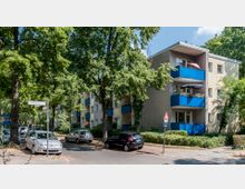 Mehrfamilienhaus in einer Wohngegend, umgeben von großen Bäumen und parkenden Autos entlang der Straße. Das Gebäude hat blaue Balkone und ist von gepflegten Grünflächen umgeben.