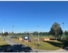 Offenes Sportgelände mit einem umzäunten Baseballfeld, mehreren Flutlichtmasten und kleinen Gebäuden am Rand. Im Hintergrund sind Bäume und ein hoher, schlanker Turm sichtbar, der von einer klaren, blauen Himmelkulisse umgeben ist.